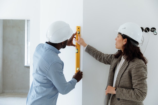 Two people in white hard hats holding a yellow level against a white wall